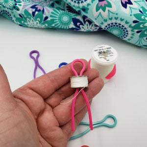 May include: A pink plastic bobbin holder with a white thread spool. The bobbin holder is shaped like a person with arms and legs. The bobbin holder is being held in a hand. There are other bobbin holders in the background, including a purple one and a blue one.