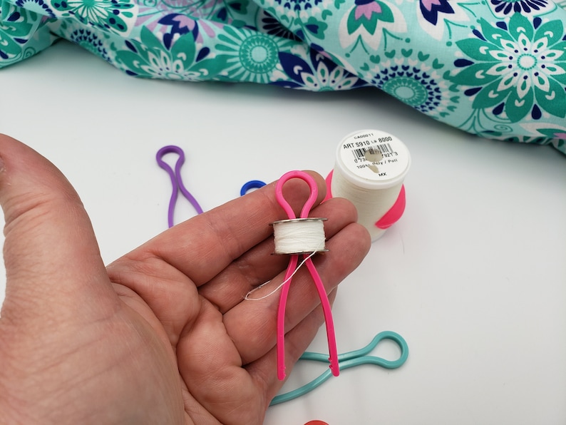 May include: A pink plastic bobbin holder with a white thread spool. The bobbin holder is shaped like a person with arms and legs. The bobbin holder is being held in a hand. There are other bobbin holders in the background, including a purple one and a blue one.
