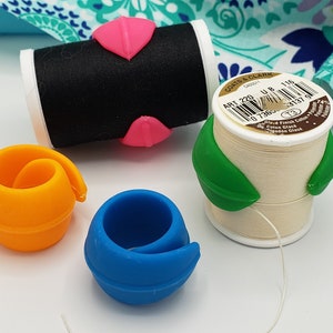 May include: Three colorful thread spool holders, one orange, one blue, and one green, with a white thread spool with a brown label that reads "COATS & CLARK ART 220 UB 116 0 73653 3137 9 (T3) Glacé Finish Cotton Coton Glacé Algódon Glacé", and a black thread spool with pink accents.