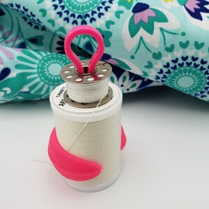 May include: A white thread spool with a pink plastic thread holder. The spool has a silver metal top with a small hole in the center. The thread holder has a loop on top and a curved shape on the bottom.