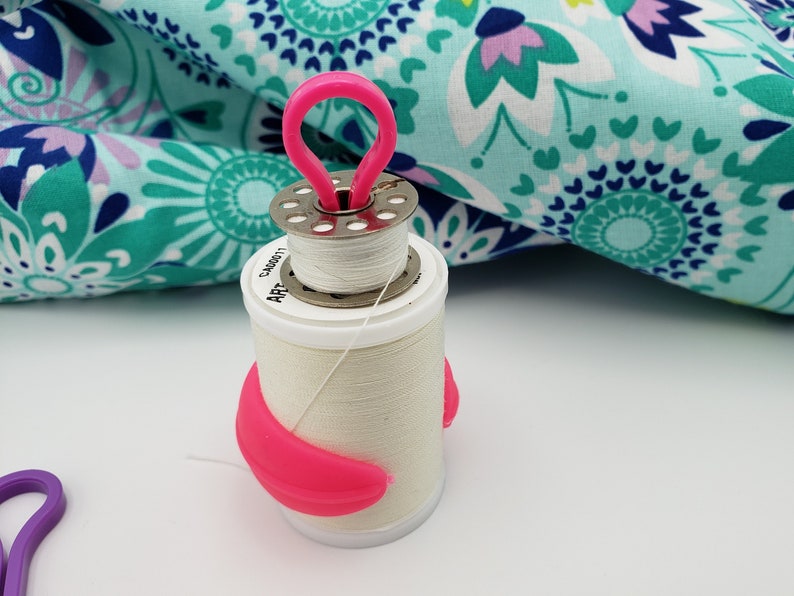 May include: A white thread spool with a pink plastic thread holder. The spool has a silver metal top with a small hole in the center. The thread holder has a loop on top and a curved shape on the bottom.
