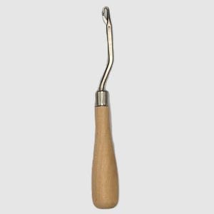 May include: A rug hooking tool with a curved, silver-colored metal hook and a light brown wooden handle. The handle is smooth and rounded, providing a comfortable grip for detailed craft work.