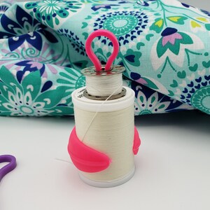 May include: A white thread spool with a pink thread holder and a pink thread guide. The spool is sitting on a white surface.