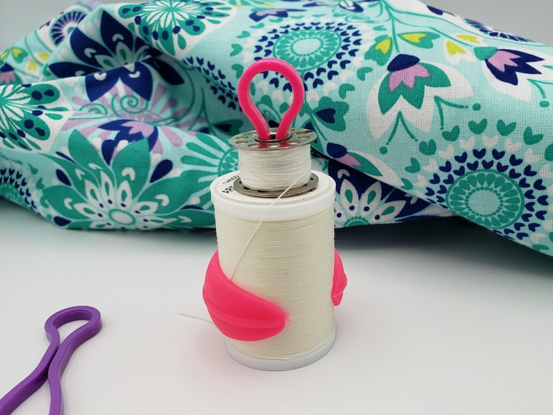 May include: A white thread spool with a pink thread holder and a pink thread guide. The spool is sitting on a white surface.