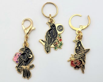 May include: Three gold-colored charms with black raven designs. One charm has a lobster clasp, another has a keyring, and the third has a lever back. Each raven is perched on a branch with floral and celestial accents in pink, red, and green.