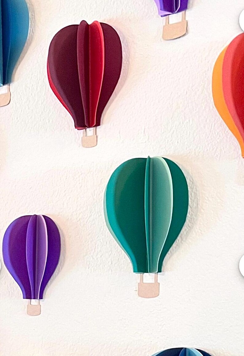 May include: A collection of colorful paper hot air balloons in various sizes and shades of blue, green, purple, red, and orange. The balloons are attached to a white wall.