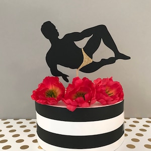 May include: Black and white silhouette cake topper of a person in a gold swimsuit. The topper is on top of a black and white striped cake with red flowers.