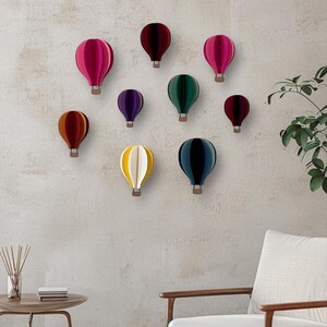 May include: A wall decor set of nine paper hot air balloons in various colors, including red, pink, purple, green, yellow, and blue. The balloons are arranged in a staggered pattern on a light gray wall.