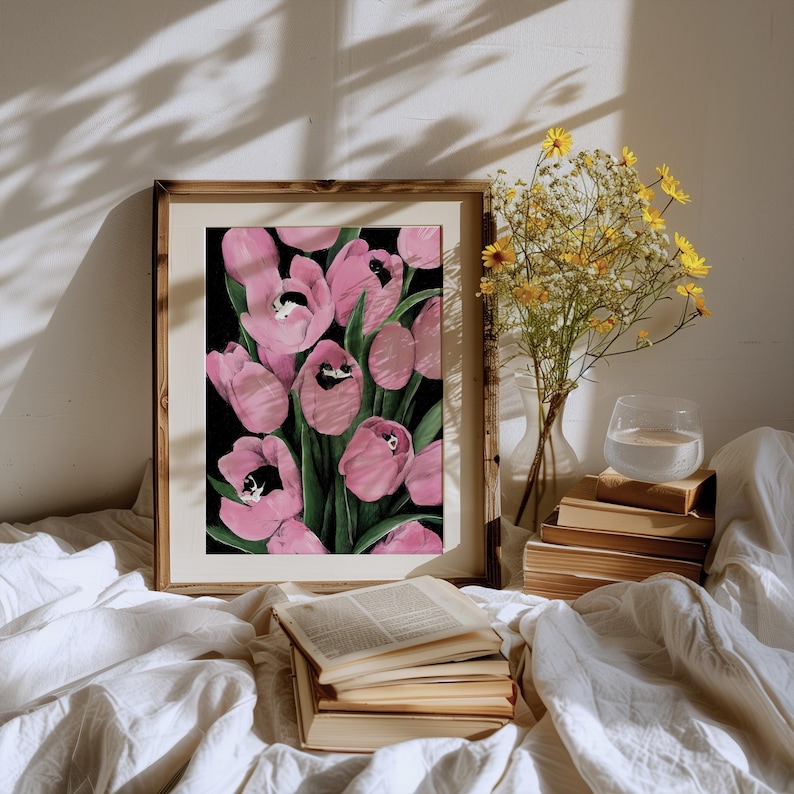 May include: A framed painting depicting pink tulips with black and white cats peeking out from inside the flowers. The painting is on a white bed with a stack of books and a vase of yellow flowers.