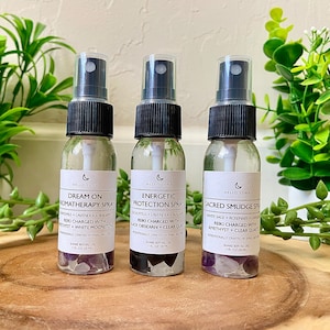May include: Three small glass spray bottles with clear liquid and crystals inside. Each bottle has a white label with the brand name "Hello Luna" and a description of the spray. The first bottle is labeled "Dream On Aromatherapy Spray" with the ingredients "Chamomile + Lavender + Bergamot". The second bottle is labeled "Energetic Protection Spray" with the ingredients "Eucalyptus + Lavender + Frankincense". The third bottle is labeled "Sacred Smudge Spray" with the ingredients "White Sage + Rosemary + Lavender".