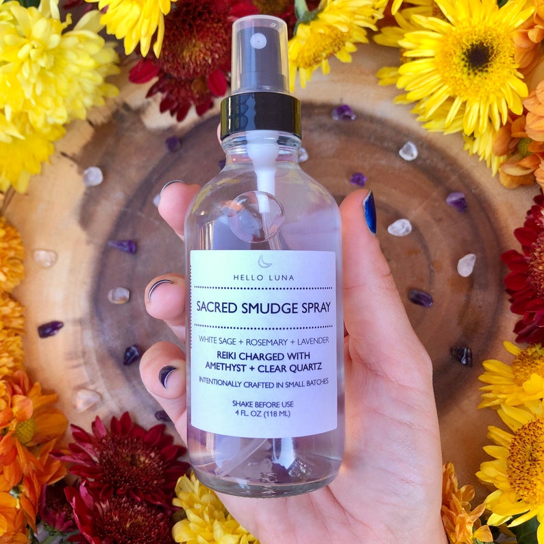 May include: A clear glass bottle with a black spray nozzle and a white label that reads "HELLO LUNA SACRED SMUDGE SPRAY WHITE SAGE + ROSEMARY + LAVENDER REIKI CHARGED WITH AMETHYST + CLEAR QUARTZ INTENTIONALLY CRAFTED IN SMALL BATCHES SHAKE BEFORE USE 4 FL OZ (118 ML)"