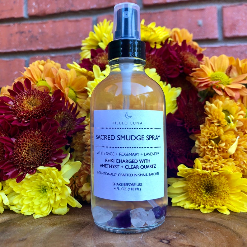 May include: A clear glass bottle with a black spray top filled with a white liquid and crystals. The bottle has a white label with the text "HELLO LUNA SACRED SMUDGE SPRAY WHITE SAGE + ROSEMARY + LAVENDER REIKI CHARGED WITH AMETHYST + CLEAR QUARTZ INTENTIONALLY CRAFTED IN SMALL BATCHES SHAKE BEFORE USE 4 FL OZ (118 ML)"
