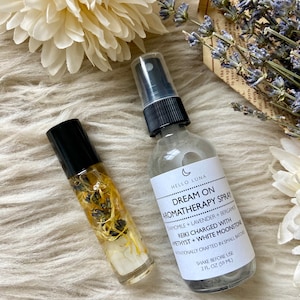 Puede incluir: Se muestran dos productos de aromaterapia: un frasco roll-on y un frasco en spray. El frasco en spray tiene una etiqueta que dice "Dream On Aromatherapy Spray". El frasco roll-on contiene flores secas y cristales, sobre una superficie blanca.