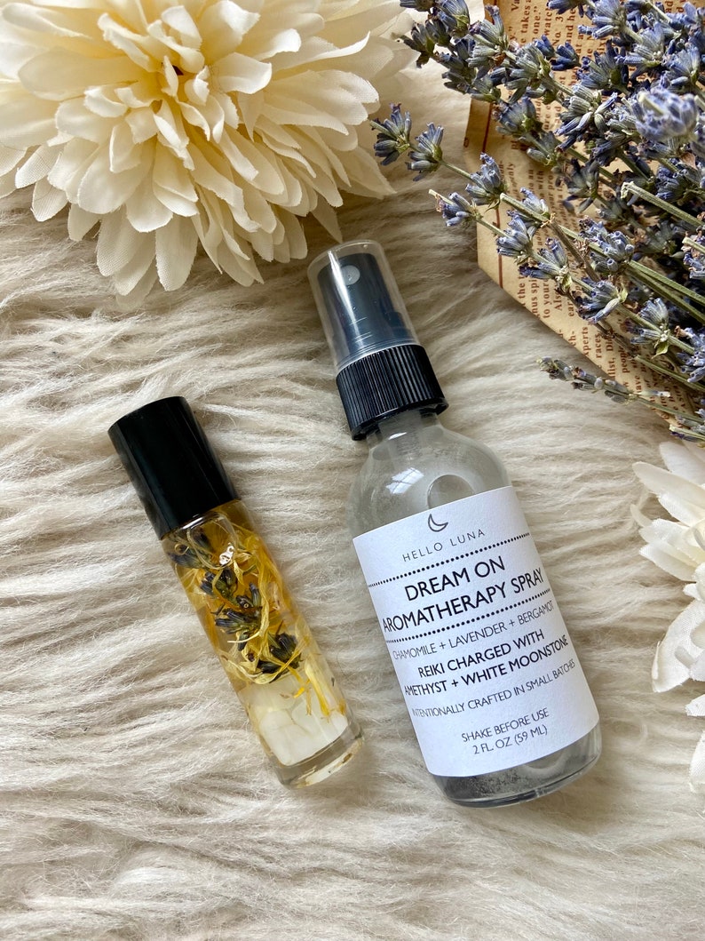 Puede incluir: Se muestran dos productos de aromaterapia: un frasco roll-on y un frasco en spray. El frasco en spray tiene una etiqueta que dice "Dream On Aromatherapy Spray". El frasco roll-on contiene flores secas y cristales, sobre una superficie blanca.