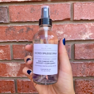 May include: A clear glass bottle with a black spray top and a white label that reads "HELLO LUNA SACRED SMUDGE SPRAY WHITE SAGE + ROSEMARY + LAVENDER REIKI CHARGED WITH AMETHYST + CLEAR QUARTZ INTENTIONALLY CRAFTED IN SMALL BATCHES SHAKE BEFORE USE 4 FL OZ (118 ML)". The bottle contains clear liquid and purple and clear crystals.
