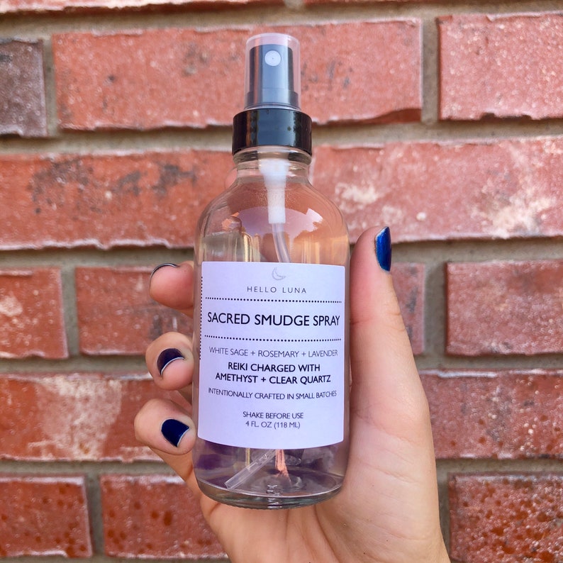 May include: A clear glass bottle with a black spray top and a white label that reads "HELLO LUNA SACRED SMUDGE SPRAY WHITE SAGE + ROSEMARY + LAVENDER REIKI CHARGED WITH AMETHYST + CLEAR QUARTZ INTENTIONALLY CRAFTED IN SMALL BATCHES SHAKE BEFORE USE 4 FL OZ (118 ML)". The bottle contains clear liquid and purple and clear crystals.
