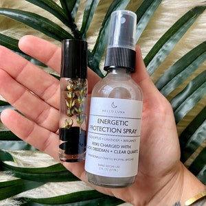 May include: A clear glass bottle with a black cap and a white label that reads "HELLO LUNA ENERGETIC PROTECTION SPRAY EUCALYPTUS + LAVENDER + BERGAMOT REIKI CHARGED WITH BLACK OBSIDIAN + CLEAR QUARTZ INTENTIONALLY CRAFTED IN SMALL BATCHES SHAKE BEFORE USE 2 FL OZ (59 ML)". The bottle is in a hand with a tattoo that reads "Live".