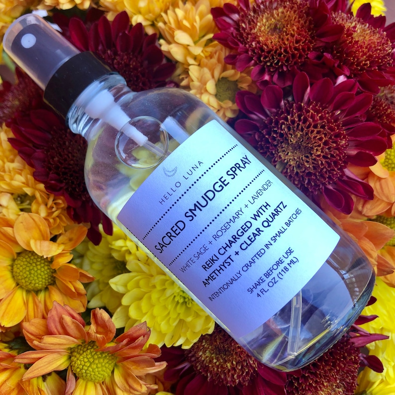 May include: A clear glass bottle with a black spray nozzle, labeled "HELLO LUNA SACRED SMUDGE SPRAY" with the ingredients "WHITE SAGE + ROSEMARY + LAVENDER" and "REIKI CHARGED WITH AMETHYST + CLEAR QUARTZ". The bottle is surrounded by a bouquet of colorful flowers.