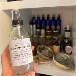 May include: A clear glass bottle with a black spray top and a white label that reads "HELLO LUNA SACRED SMUDGE SPRAY WHITE SAGE + ROSEMARY + LAVENDER REIKI CHARGED WITH AMETHYST + CLEAR QUARTZ INTENTIONALLY CRAFTED IN SMALL BATCHES SHAKE BEFORE USE 4 FL OZ (118 ML)"