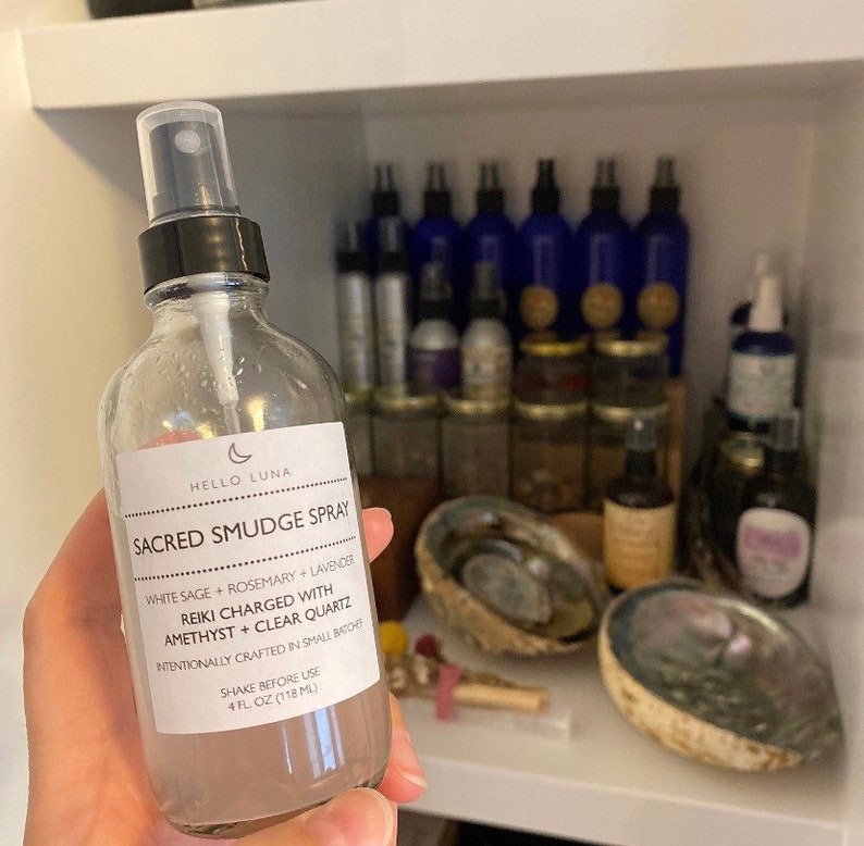 May include: A clear glass bottle with a black spray top and a white label that reads "HELLO LUNA SACRED SMUDGE SPRAY WHITE SAGE + ROSEMARY + LAVENDER REIKI CHARGED WITH AMETHYST + CLEAR QUARTZ INTENTIONALLY CRAFTED IN SMALL BATCHES SHAKE BEFORE USE 4 FL OZ (118 ML)"