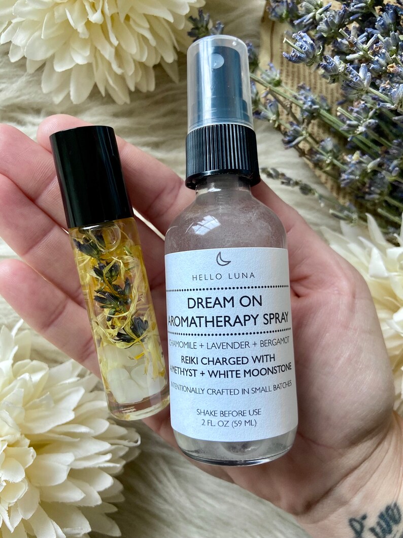 Puede incluir: Se muestran dos productos de aromaterapia: un frasco roll-on y un frasco en spray. El roll-on contiene elementos bot&aacute;nicos visibles. El frasco en spray, etiquetado como "Dream On Aromatherapy Spray", es de 59 ml y contiene manzanilla y lavanda.