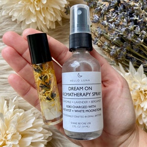Puede incluir: Una mano sostiene un frasco roll-on y un frasco de spray de productos de aromaterapia. El frasco de spray tiene una tapa negra y una etiqueta que dice "Dream On Aromatherapy Spray". El frasco roll-on contiene flores secas y cristales.