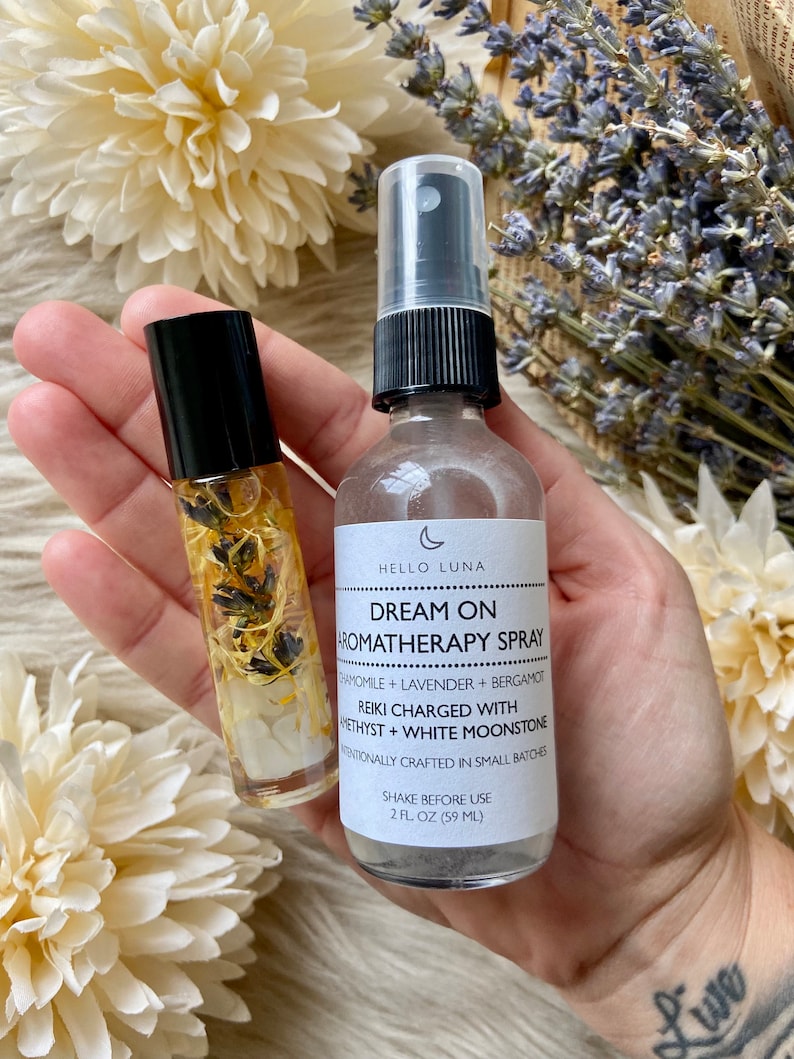 Puede incluir: Una mano sostiene un frasco roll-on y un frasco de spray de productos de aromaterapia. El frasco de spray tiene una tapa negra y una etiqueta que dice "Dream On Aromatherapy Spray". El frasco roll-on contiene flores secas y cristales.