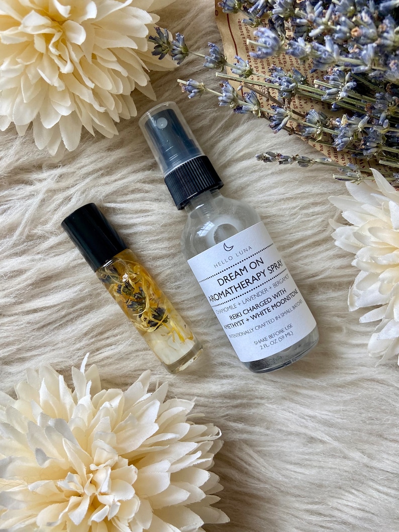 Puede incluir: Productos de aromaterapia, que incluyen un frasco rociador y un frasco roll-on, se exhiben sobre una superficie blanca texturizada. El frasco rociador tiene una tapa negra y una etiqueta que dice "Dream On Aromatherapy Spray". El frasco roll-on contiene elementos bot&aacute;nicos visibles.