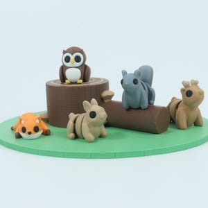 Puede incluir: Un conjunto de cinco figuritas de animales del bosque, que incluyen un búho, una ardilla, un ciervo, un conejo y un zorro, están dispuestos sobre un tocón de árbol y un tronco marrones. Las figuritas están hechas de resina pintada de marrón, gris y naranja.