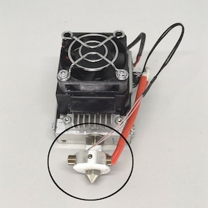 May include: A close-up of a 3D printer hot end with a silver nozzle and a black fan. The hot end is attached to a silver heatsink and has a red and white wire attached to it.