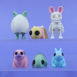以下が含まれることがあります： 6体の小さなカラフルな3Dプリントされたフィギュアのセット。フィギュアは、ウサギ、パグ、カメ、ミツバチなど、さまざまな動物の形をしています。フィギュアはすべて透明なアクリル製の棚の上に立っています。
