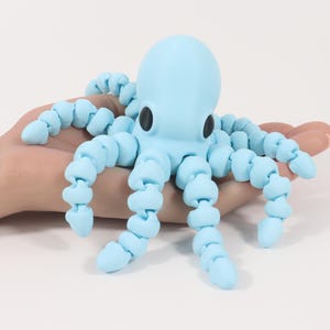 May include: A light blue octopus toy with segmented tentacles and black eyes. The toy is held in a person's hand against a white background. The octopus is a whimsical, playful design.