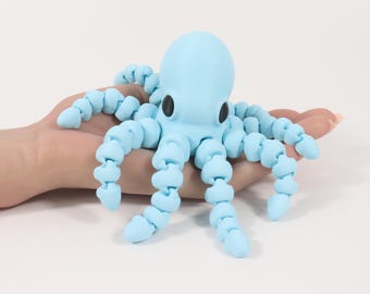 May include: A light blue octopus toy with segmented tentacles and black eyes. The toy is held in a person's hand against a white background. The octopus is a whimsical, playful design.
