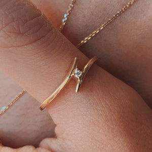 May include: A close-up of a hand wearing a delicate gold ring with a small diamond. The ring is on the index finger and is set against a backdrop of a gold chain necklace.