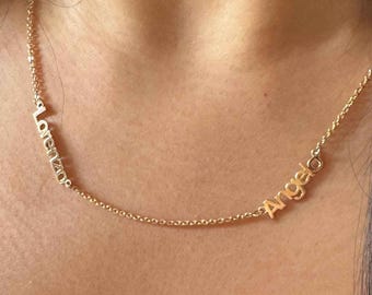 May include: A gold-colored necklace with the names "Lorenzo" and "Angelos" spelled out in delicate, cursive lettering. The necklace is worn around the neck, with the names positioned across the collarbone.