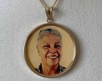 May include: A gold-colored pendant necklace featuring a circular pendant with a portrait of a smiling person. The pendant is suspended from a delicate gold chain, displayed on a mannequin.