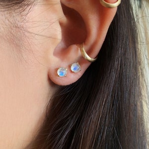 May include: A pair of gold stud earrings with iridescent moonstone gemstones. The earrings are worn in the lobes of a person's ears.