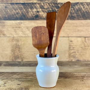 May include: Three wooden kitchen utensils, a spatula, a spoon, and a turner, are standing upright in a white ceramic vase. The vase has a rounded, bulbous shape and a smooth, glossy finish.