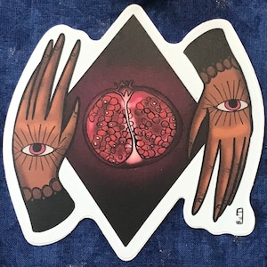 May include: Two hands with eyes on the palms, holding a red pomegranate open, against a dark purple background. The hands are surrounded by a white diamond shape.