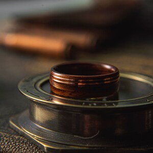 Op de afbeelding: Een houten ring met drie dunne koperen banden. De ring ligt op een ronde, messingkleurige standaard.