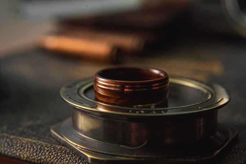 Op de afbeelding: Een houten ring met drie dunne koperen banden. De ring ligt op een ronde, messingkleurige standaard.