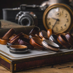 May include: A collection of wooden rings with various wood grain patterns and colors, including brown, black, and white. The rings are arranged on a white surface with a gold trim.