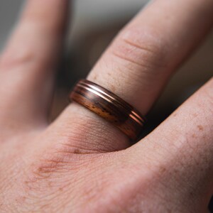 Op de afbeelding: Een houten ring met twee dunne koperen banden. De ring wordt gedragen aan een vinger.