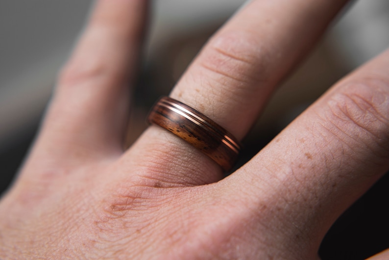 Op de afbeelding: Een houten ring met twee dunne koperen banden. De ring wordt gedragen aan een vinger.