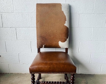 May include: A wooden chair with a brown leather seat and a cowhide back. The chair has dark wood legs with a twisted design and brass nailhead trim. The back of the chair is a mix of brown and white cowhide.