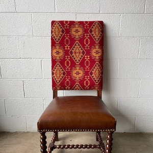 May include: A wooden chair with a brown leather seat and a red, yellow, and orange patterned back. The chair has a twisted design on the legs.