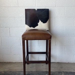 May include: A brown leather bar stool with a black and white cowhide seat back.