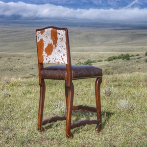 May include: A brown wooden bar stool with a cowhide seat and back. The stool is set against a backdrop of a grassy field and mountains.