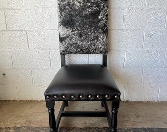 May include: Black leather seat chair with a black and white cowhide back. The chair has a black wooden frame and brass nailhead trim around the seat. The chair is a decorative piece.