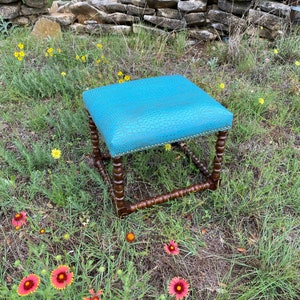 May include: A turquoise blue square ottoman with a faux crocodile skin pattern. The ottoman has a wooden base with turned legs and decorative nailheads around the top edge.